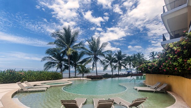 A circular pool with lounge chairs, palm trees, and a view of the ocean. - Vistas Las Palmas by Dream Makers (Jacó)