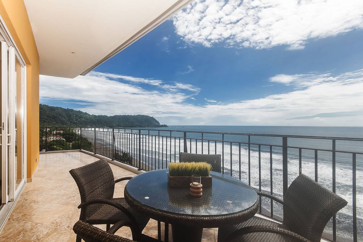Un balcón con una mesa redonda y sillas de mimbre, con vistas a la costa y un cielo despejado.