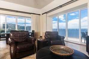 Un vaste salon doté de canapés en cuir, d’un coin repas et de grandes fenêtres offrant une vue sur la mer et les collines.