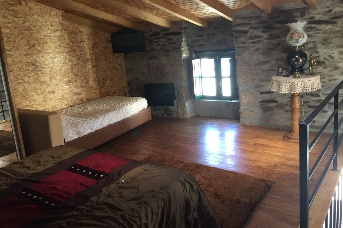 A loft bedroom with a loft bed, a stone wall, a wooden ceiling, a small window, and a decorative table.