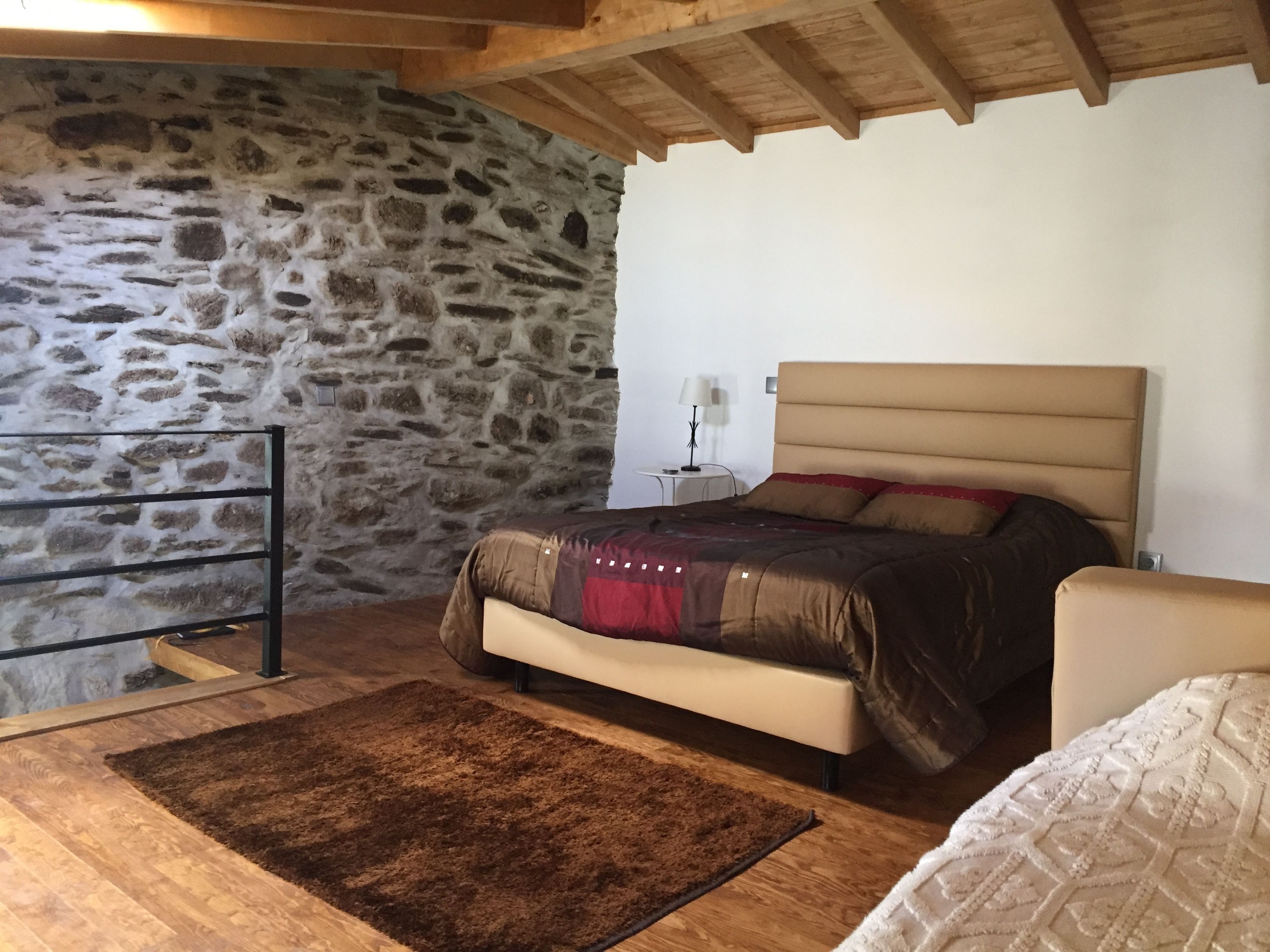 Une chambre avec un mur en pierre, un lit avec une tête de lit, une table de chevet, une lampe et un plancher en bois.