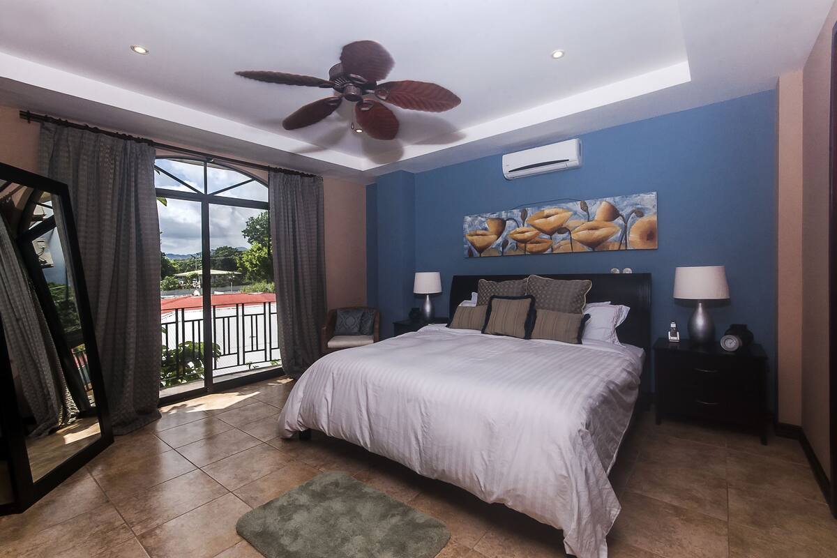 A bedroom with a large bed, a ceiling fan, a nightstand with a lamp, and a painting on the wall.