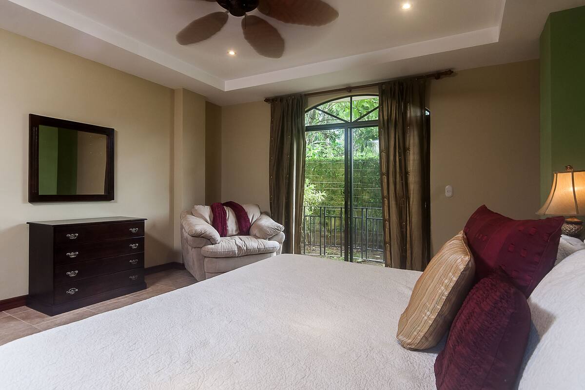 A bedroom with a large bed, a dresser, a chair, a ceiling fan, and a window with curtains.