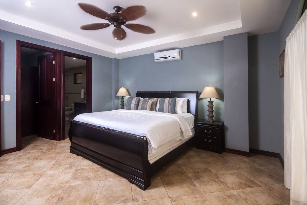 A bedroom with a dark wooden bed, two bedside tables with lamps, a ceiling fan, and an air conditioning unit.