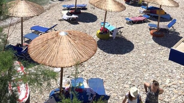 En strand med parasoller og liggestole, klippefremspring og klart blåt vand.