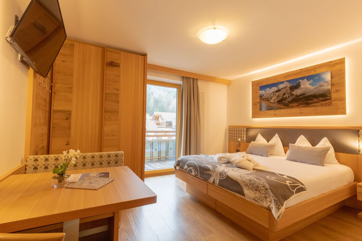 Una habitación de hotel con una cama de madera, un televisor montado en la pared, una mesa de comedor con un florero y un amplio ventanal con vistas al exterior.