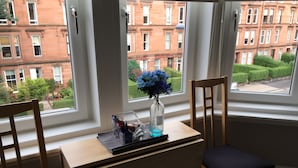 Une pièce avec un bureau et deux chaises, un vase rempli de fleurs, et une vue sur des bâtiments en briques rouges et un jardin verdoyant, le tout visible à travers de grandes fenêtres.