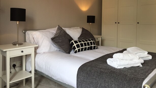 Un lit bien fait, avec du linge de lit blanc et un plaid gris, une table de chevet avec une lampe et une armoire à portes.