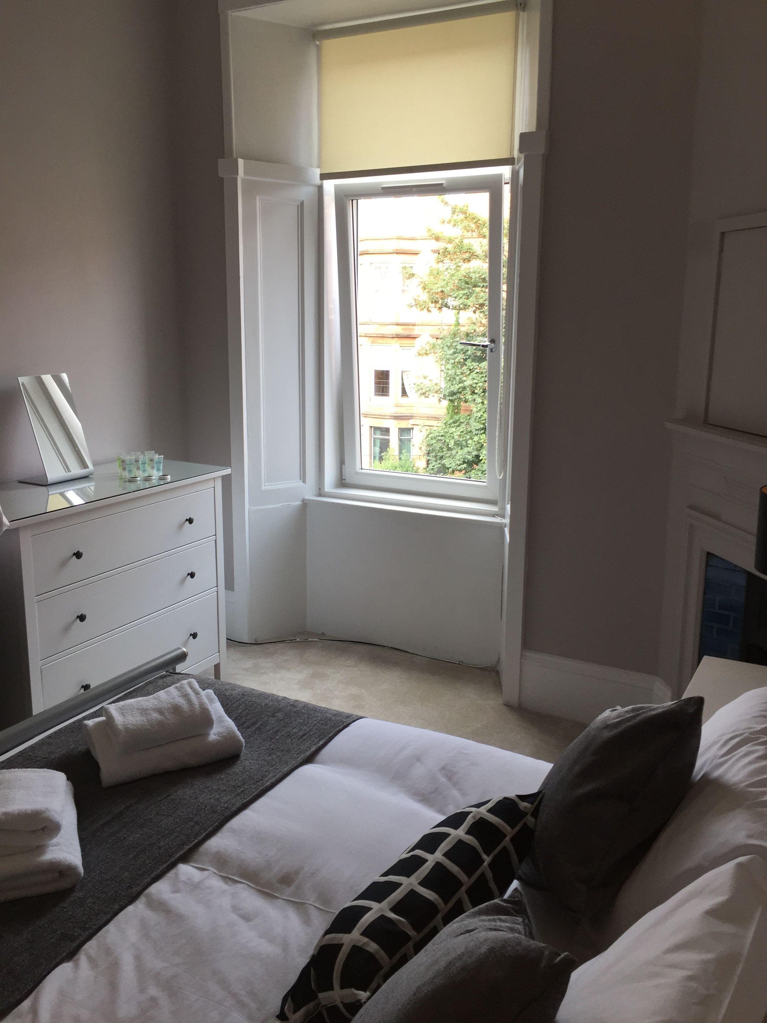 A bedroom with a bed, a dresser, a window, and a view of buildings outside.