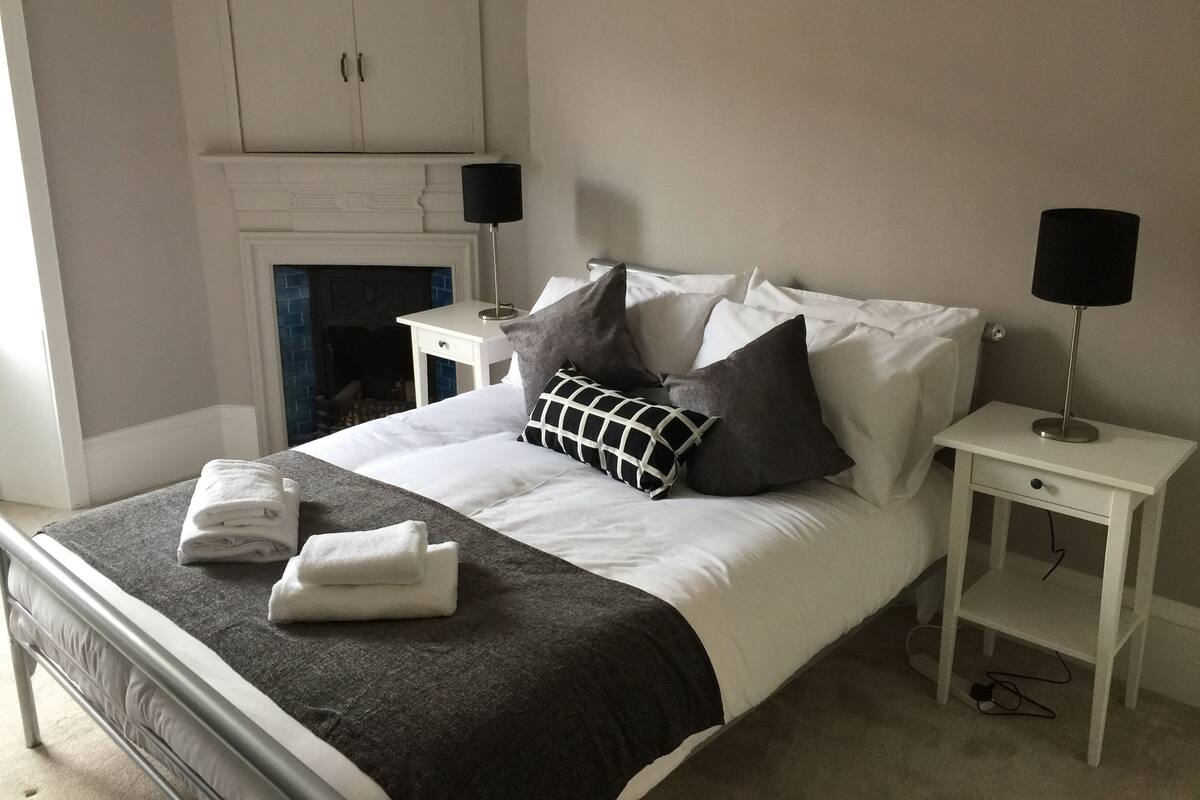 Una cama bien hecha con un cojín a cuadros blanco y negro, almohadas blancas y toallas dobladas sobre un edredón gris oscuro. Hay dos mesitas de noche con lámparas, un armario blanco y una chimenea de ladrillo azul.