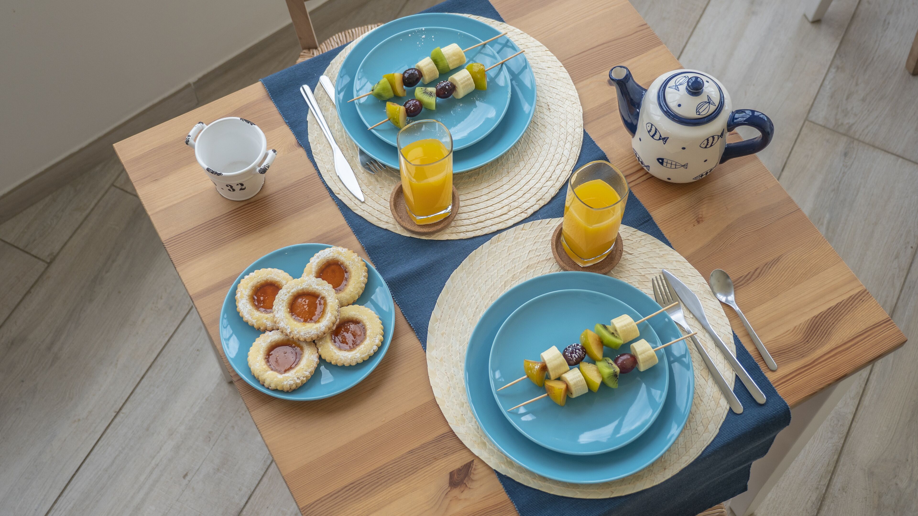 Un desayuno con brochetas de fruta, un vaso de jugo de naranja y una tetera sobre una mesa de madera.