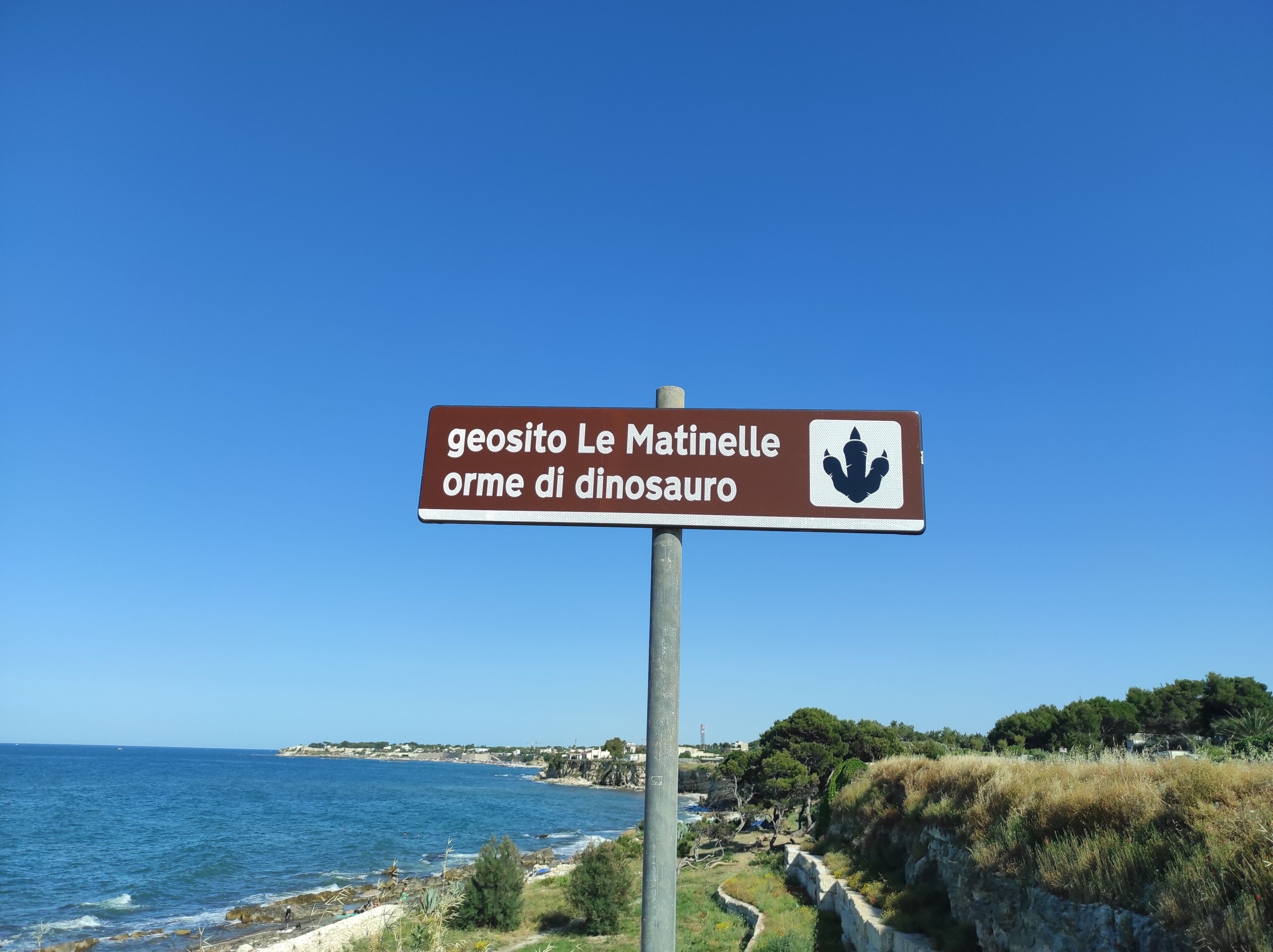 Een bord met tekst in het Italiaans, een kustlandschap en de zee.