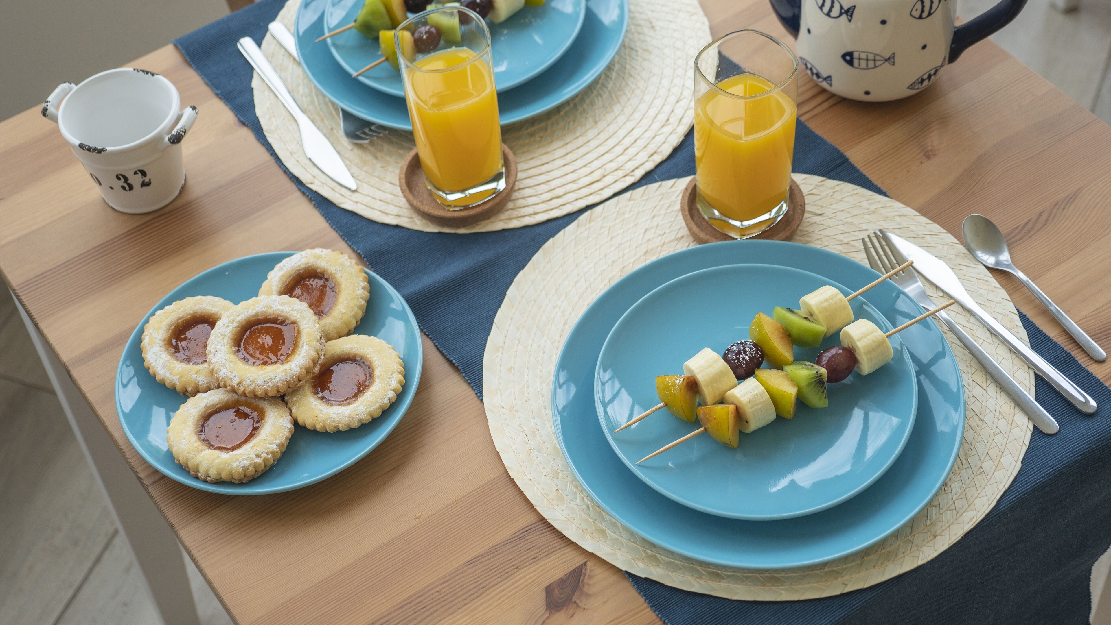 A breakfast table with plates of fruit skewers, a plate of jam-filled cookies, and glasses of orange juice.