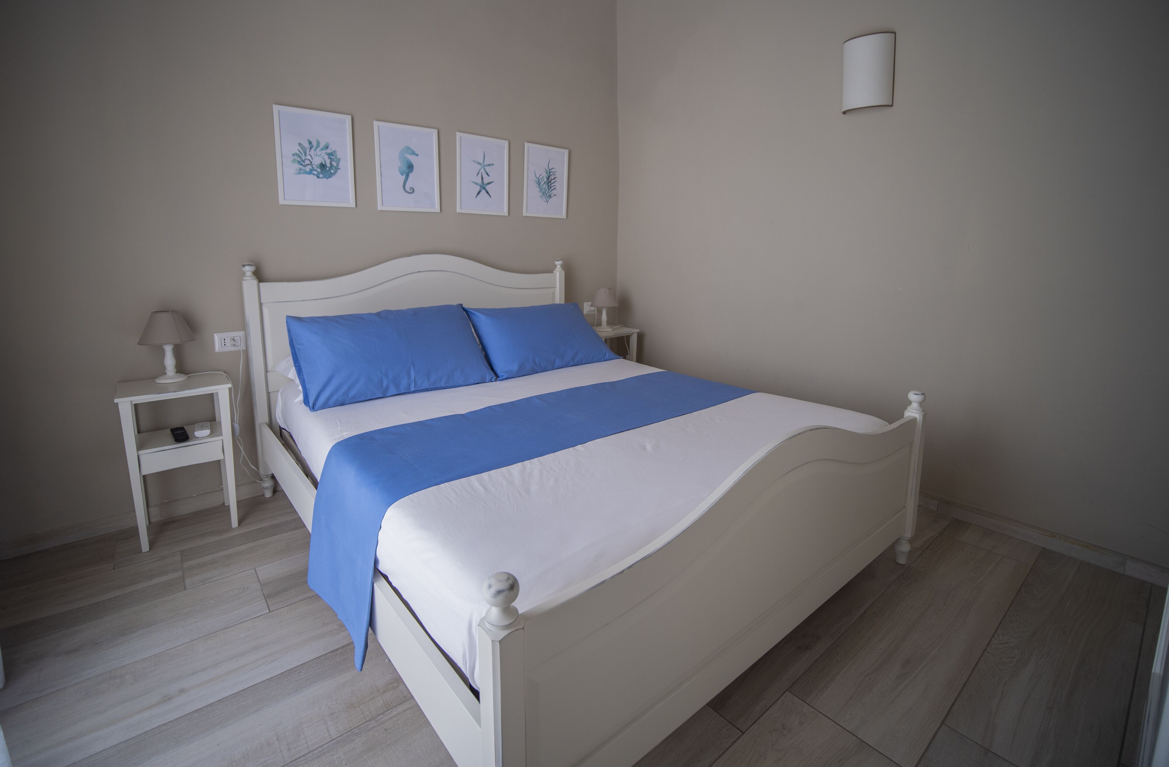 A bedroom with a white bed, blue pillows, and a white nightstand with a lamp. There are three framed pictures on the wall above the bed.