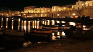 Un puerto nocturno con barcos amarrados, edificios iluminados y un muro de piedra.