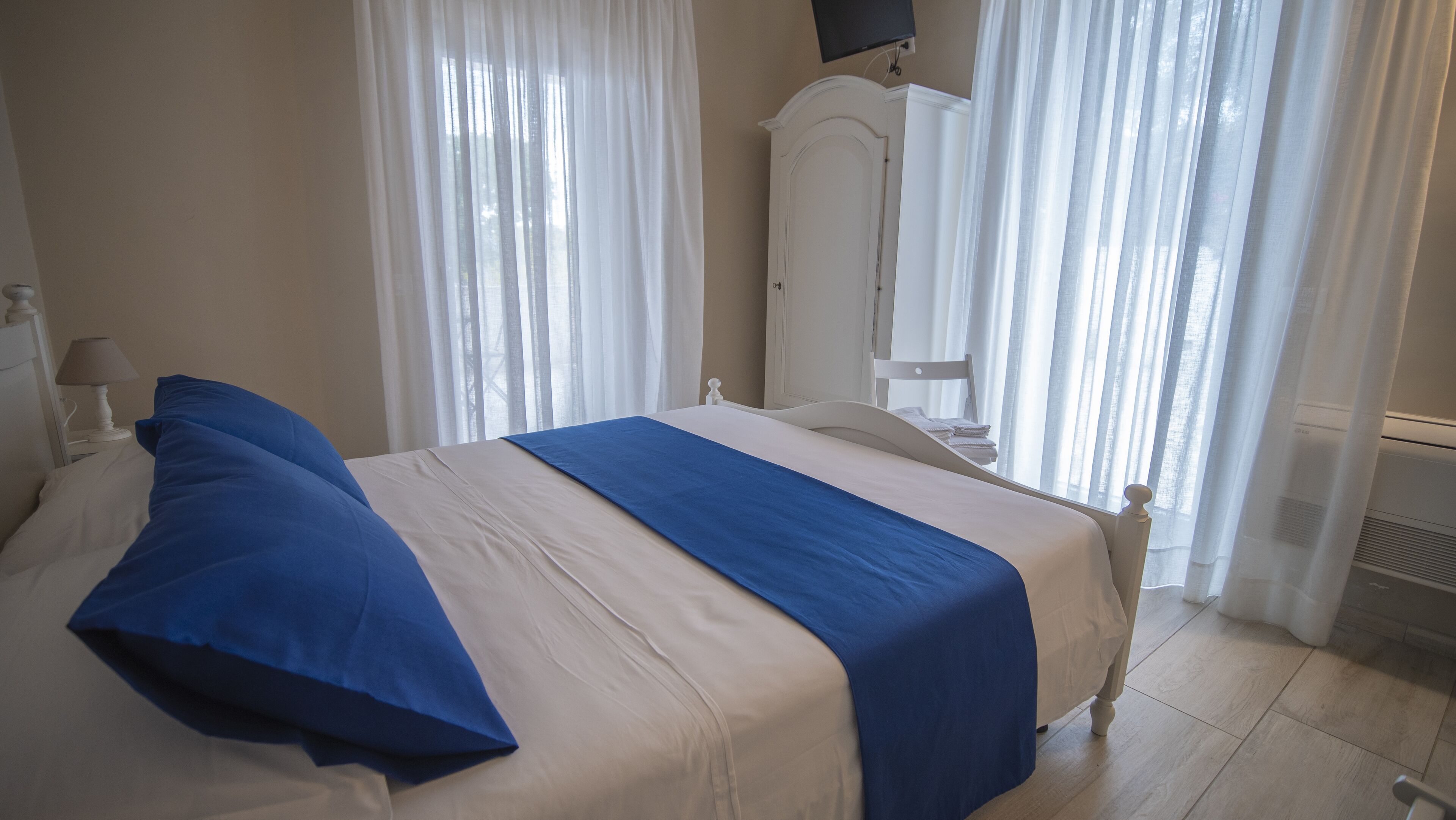 Cama bem arrumada com detalhe azul, cortinas brancas, guarda-roupa branco e televisão de tela plana fixada na parede.