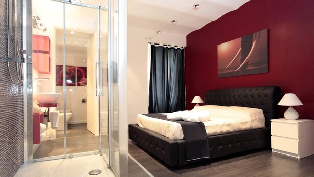 A modern bedroom with a glass shower enclosure, a large bed with a black headboard, and a red accent wall. - Rome Services Borgo Suites (Rome)