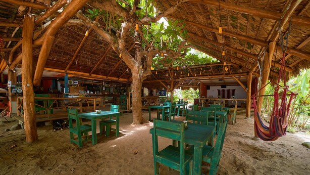 An outdoor dining area with wooden tables and chairs, a thatched roof, and a hammock. - Playa 506 Beachfront Hostel (Cahuita)