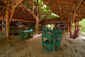 Un área de comedor al aire libre con mesas y sillas de madera, techo de paja y una hamaca.