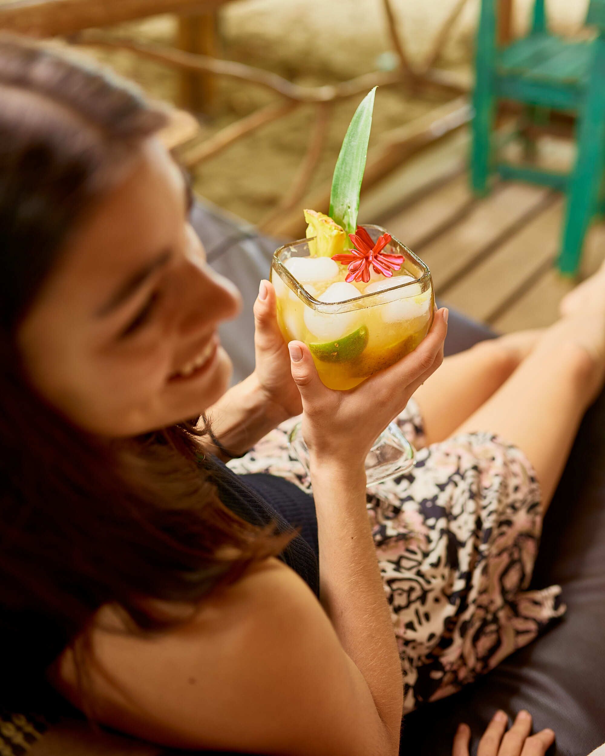 En person som håller i en cocktail med lime och en dekorativ blomma, sittande på ett trädäck.