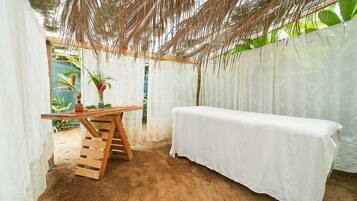 A covered outdoor area with a wooden table and a white tablecloth, surrounded by a white wall and a thatched roof.