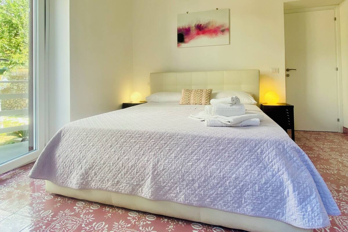 A large bed with a white quilted bedspread, two bedside lamps, and a wall-mounted abstract painting.