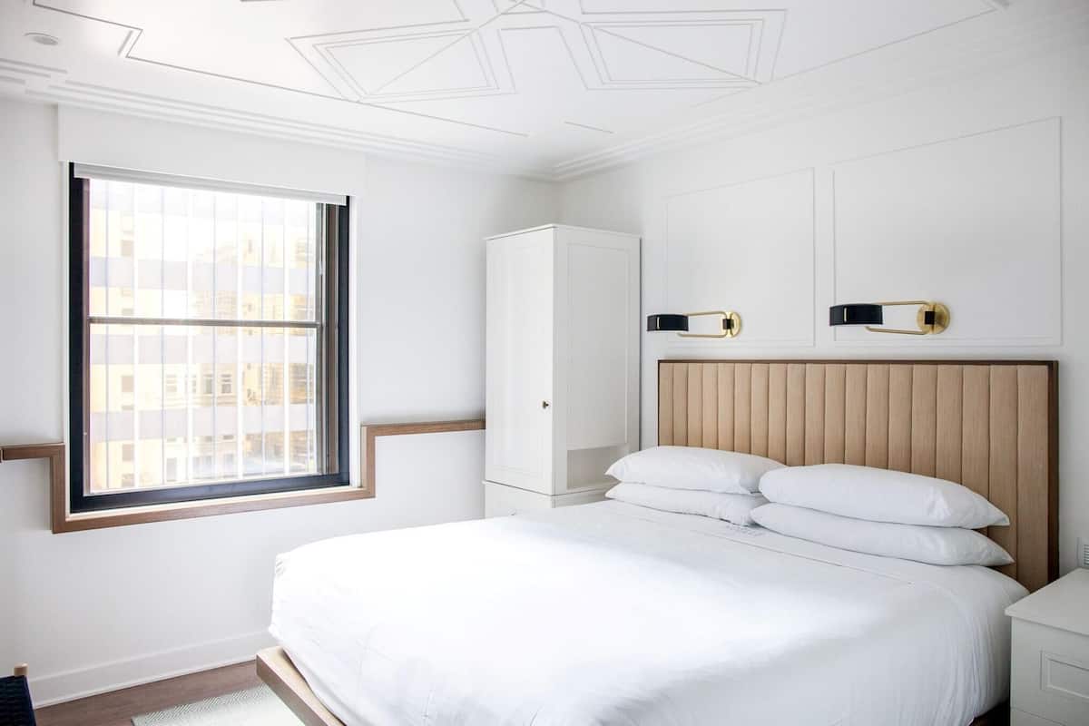 A bedroom with a large window, a bed with white linens, a wooden headboard, and a white wardrobe.