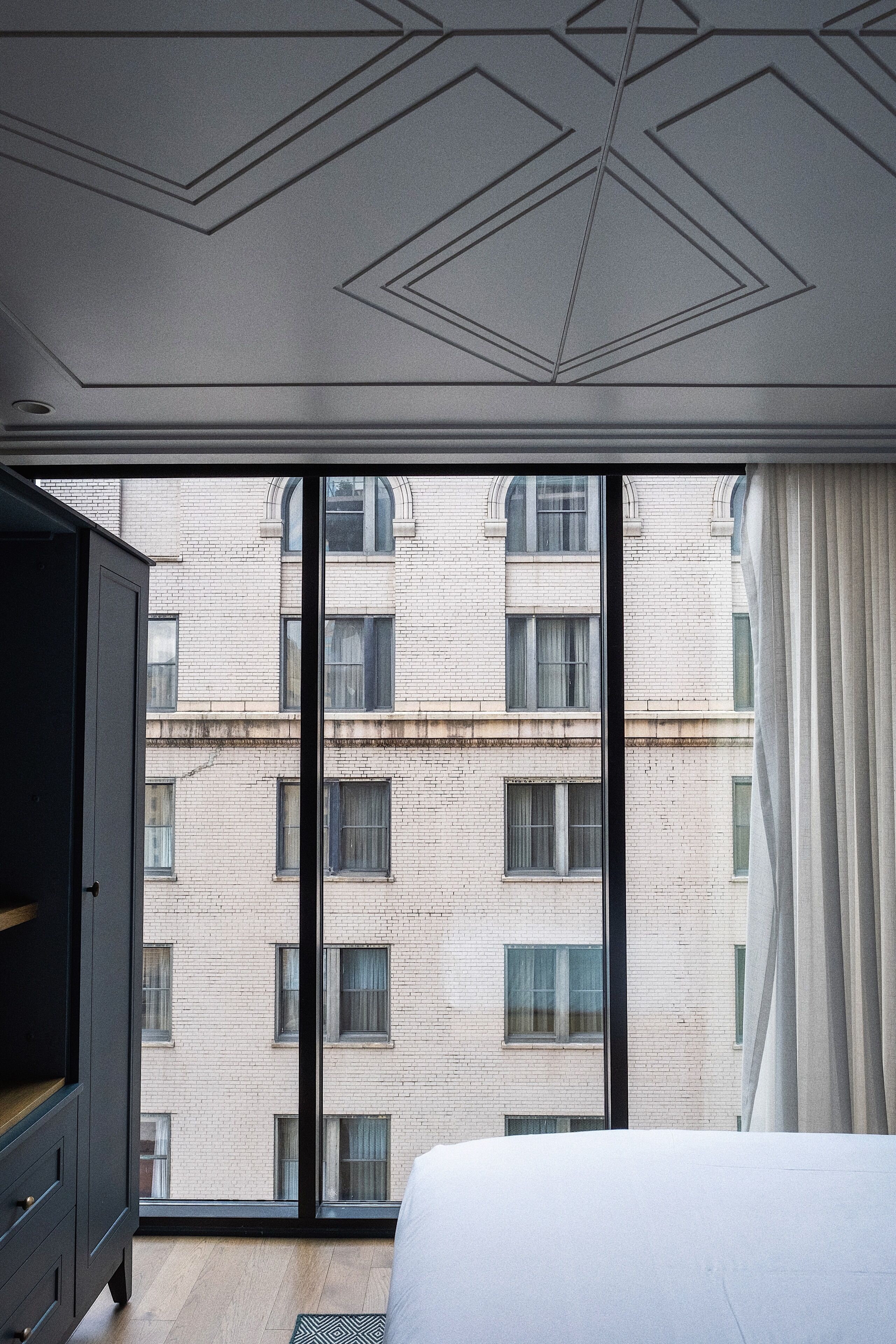 A hotel room with a large window, a bed, a wardrobe, and a geometric ceiling design.