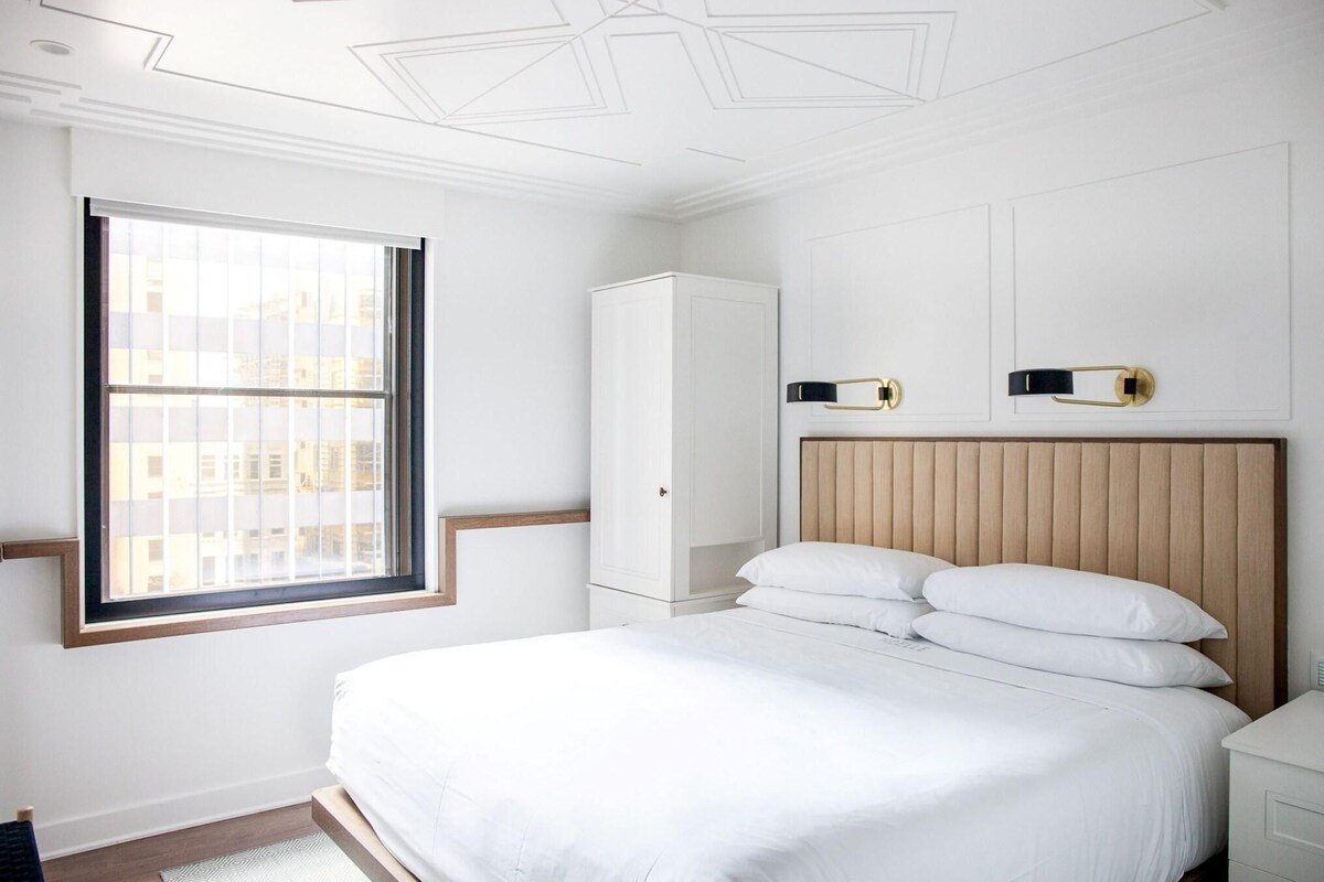 A bedroom with a large window, a bed with white linens, a wooden headboard, and a white wardrobe.
