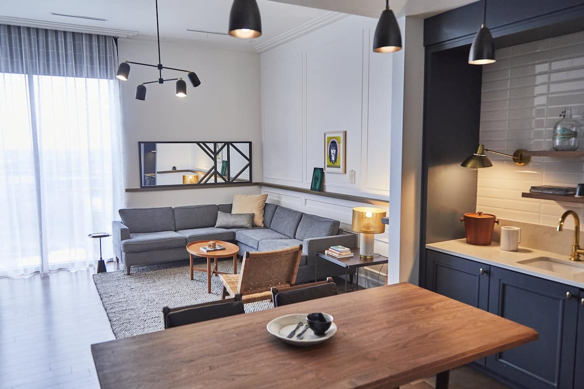 A modern living room with a sofa, coffee table, and dining table. There is a kitchen area with a sink and cabinets.