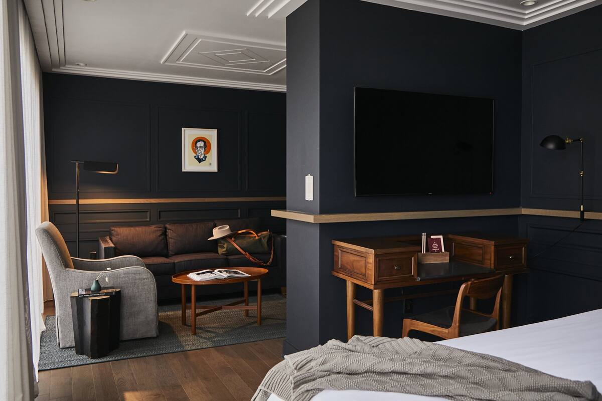 A hotel room with a dark-colored wall, a flat-screen TV mounted on the wall, a wooden desk with a chair, a sofa, a small side table, and a lamp.