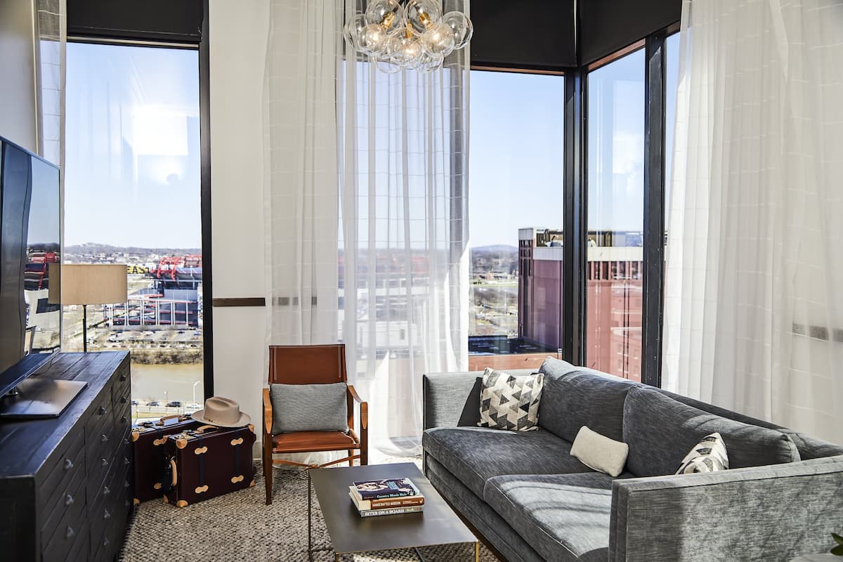 A modern living room with a grey sofa, a wooden chair, a coffee table, and a TV on a black cabinet. There is a large window with curtains, a chandelier, and a view of a cityscape.