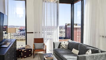 A modern living room with a grey sofa, a wooden chair, a coffee table, and a TV on a black cabinet. There is a large window with curtains, a chandelier, and a view of a cityscape.