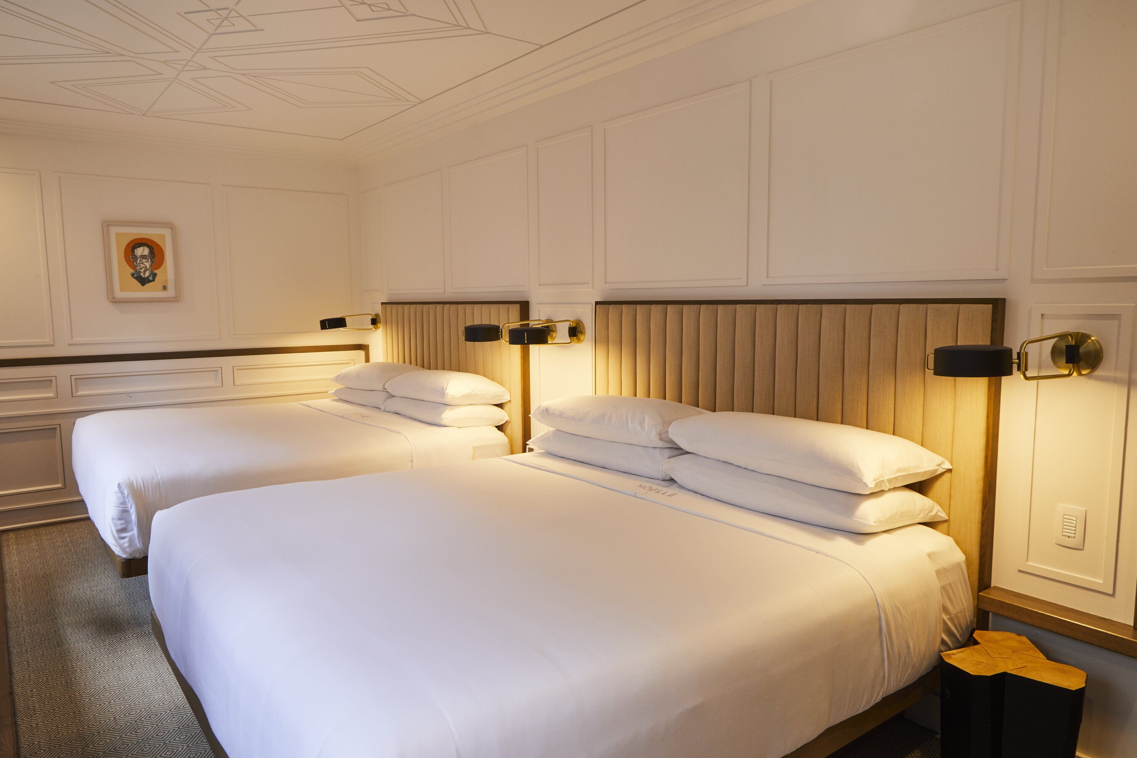 A hotel room with two beds, each with white linens and headboards, a small bedside table, and a wall-mounted light fixture.