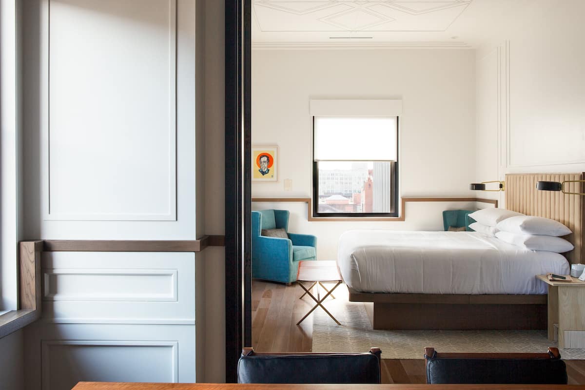A modern hotel room with a large bed, a small table, a chair, and a window with a city view.