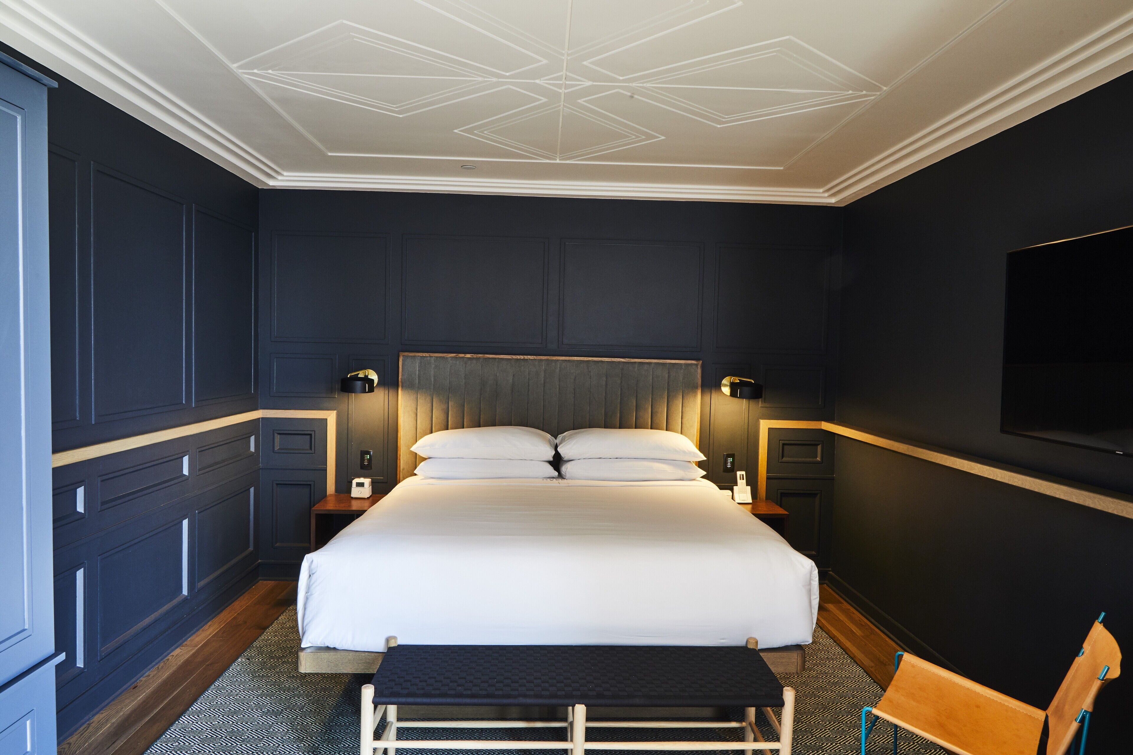Une chambre d’hôtel moderne avec un grand lit, des murs de couleur sombre et un plafond au design géométrique.