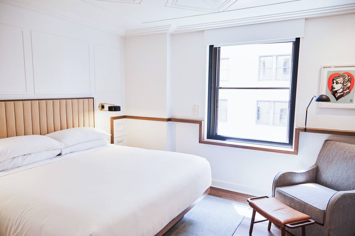 A hotel room with a large bed, a bench, a sofa, and a window with a view of buildings.