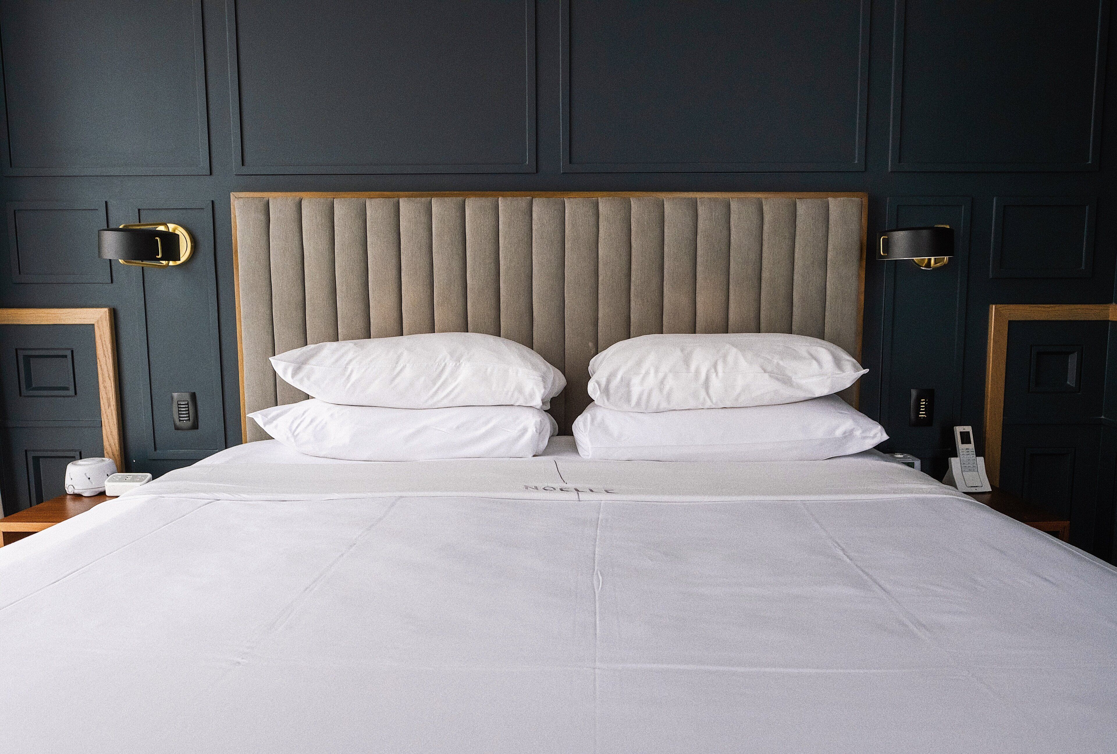 Una cama bien hecha con cabecera acolchada, ropa de cama blanca y una mesita de noche con lámpara y teléfono.
