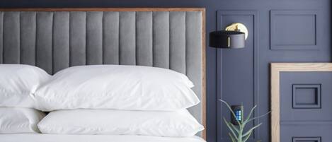 A neatly made bed with white linens and a grey upholstered headboard. A small bedside table with a potted plant and a black lamp is visible. The wall is painted in a dark blue color with paneling.