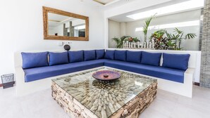 Una sala de estar moderna con un sofá seccional azul, una mesa de centro de vidrio con base de madera, un espejo grande y vistas a una pared de piedra y plantas.