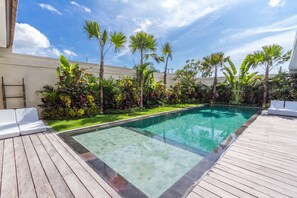 A swimming pool with a wooden deck, surrounded by tropical plants and palm trees.