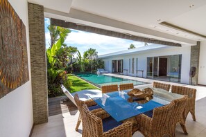 A modern outdoor dining area with a glass table and wicker chairs, overlooking a swimming pool and lush garden.