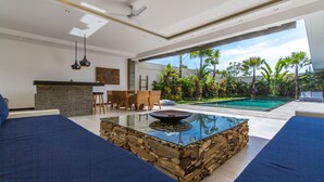 Una sala moderna con una mesa de centro de cristal, sofás azules, un comedor con mobiliario de mimbre y vistas a una piscina y plantas tropicales.