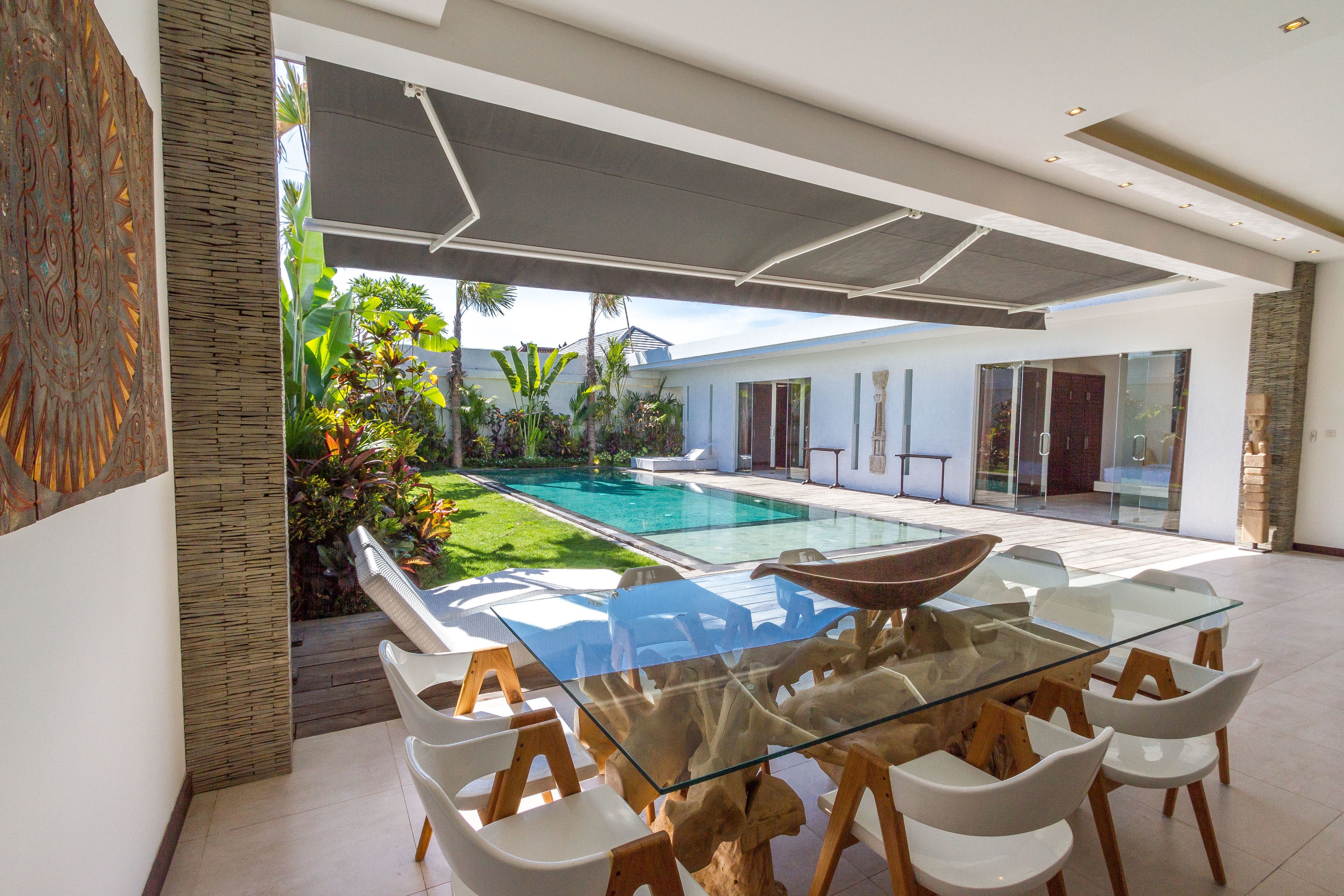 A modern outdoor dining area with a glass table and white chairs, overlooking a swimming pool and lush garden.