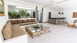 A spacious living area with a large sectional sofa, a glass coffee table, a kitchen area with bar stools, and a view of the outdoors through a large window. - Villa Nala (Seminyak)
