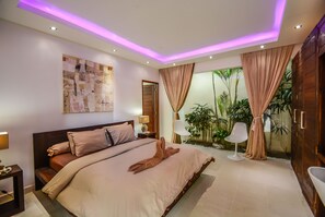 A modern bedroom with a large bed, a nightstand, a bedside table, a mirror, a window with curtains, and a view of greenery outside.