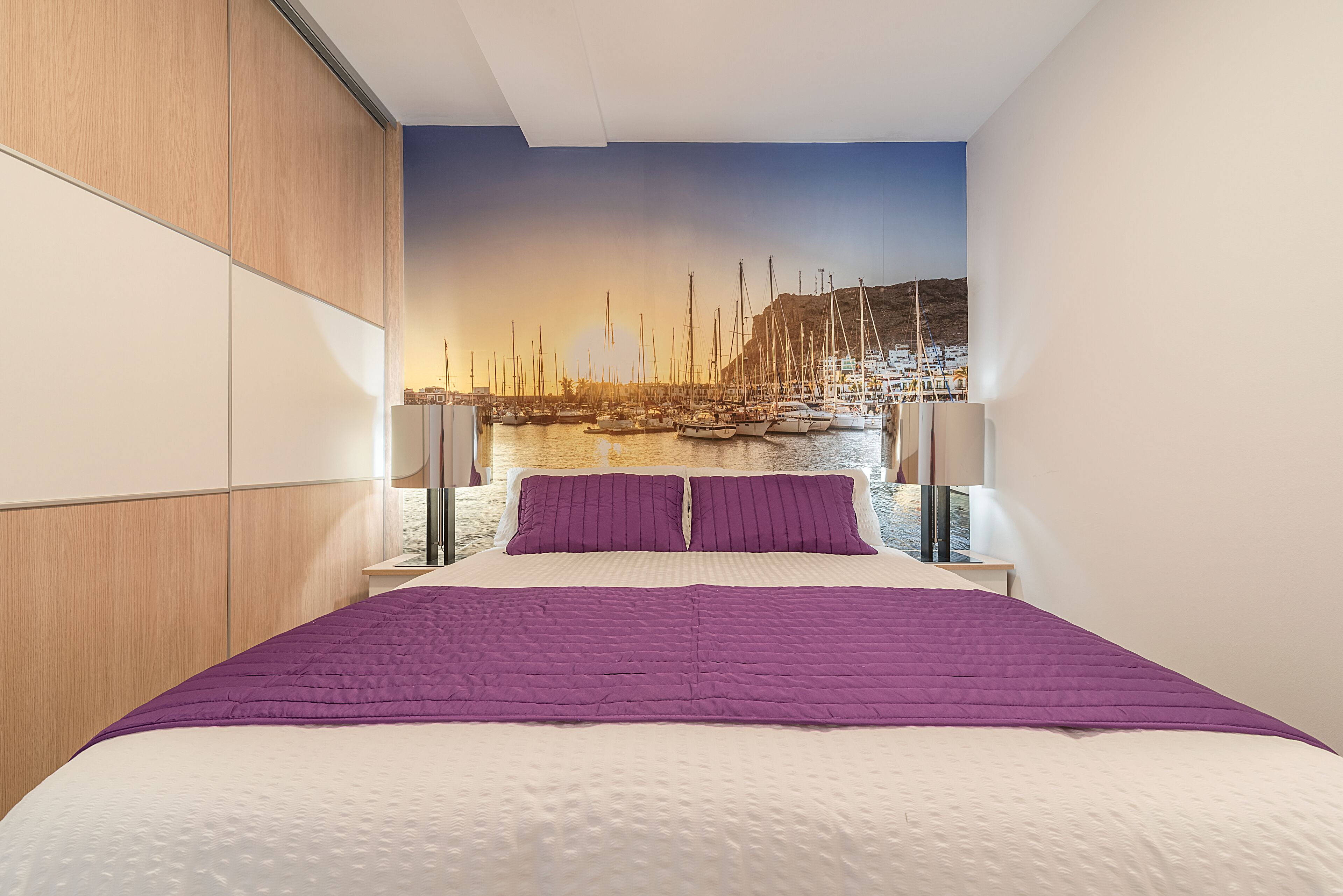 A bedroom with a large bed, a wooden wardrobe, and a mural of a marina.