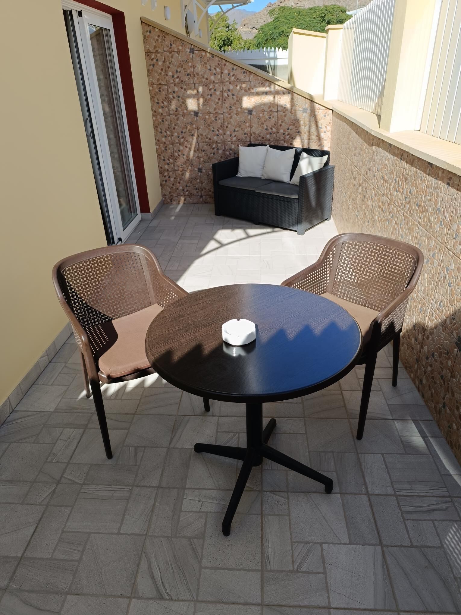 Eine Terrasse mit rundem Tisch, zwei Stühlen, einer Couch und einer Schiebetür.
