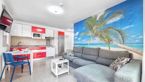 A modern living room with a grey sofa, a dining table with chairs, a kitchen area with red and white cabinets, and a large beach-themed mural on the wall. - Sol Mogan Suites (Mogan)