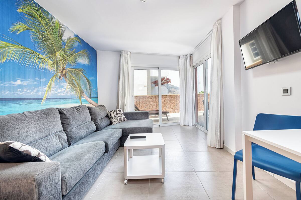 Un salon moderne avec un canapé gris, une table basse blanche, un fauteuil bleu, un téléviseur à écran plat et une grande fenêtre donnant sur la mer.